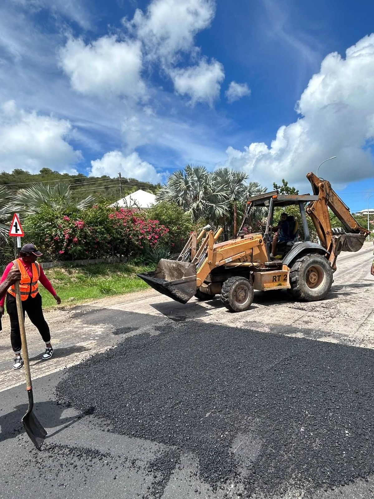 Road construction workers operate paving machinery while laying fresh asphalt along a roadway in a tropical Caribbean setting.
