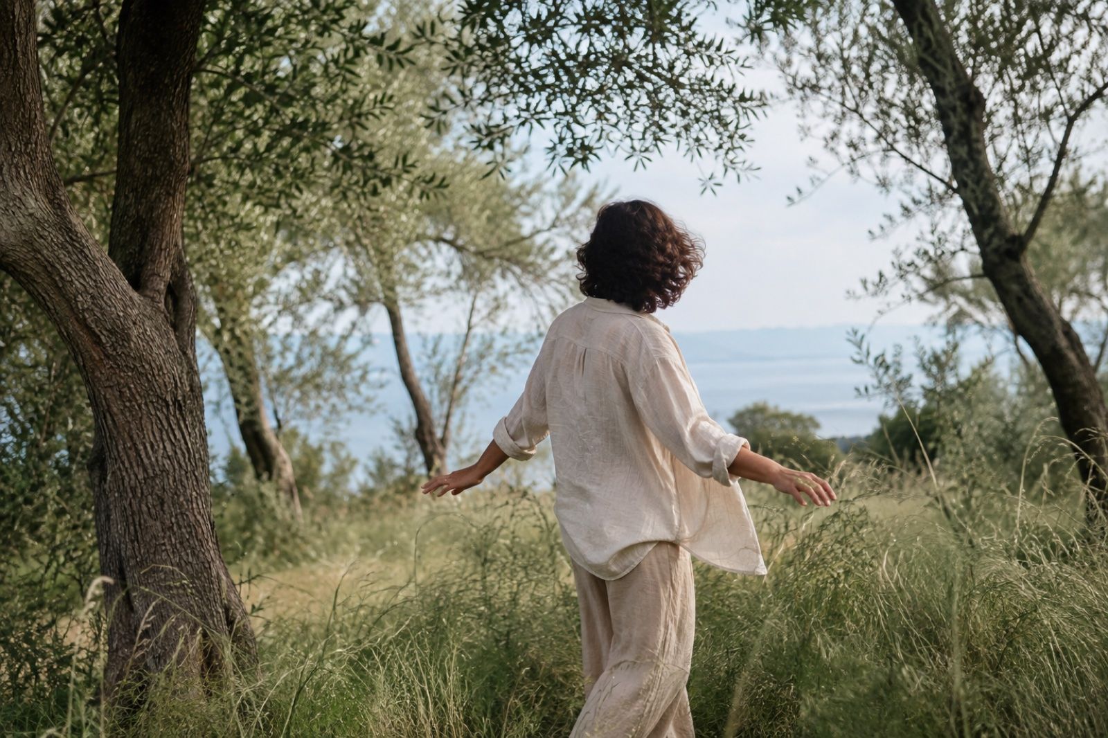 Una persona si muove lentamente tra gli ulivi e l’erba alta, vista di spalle.