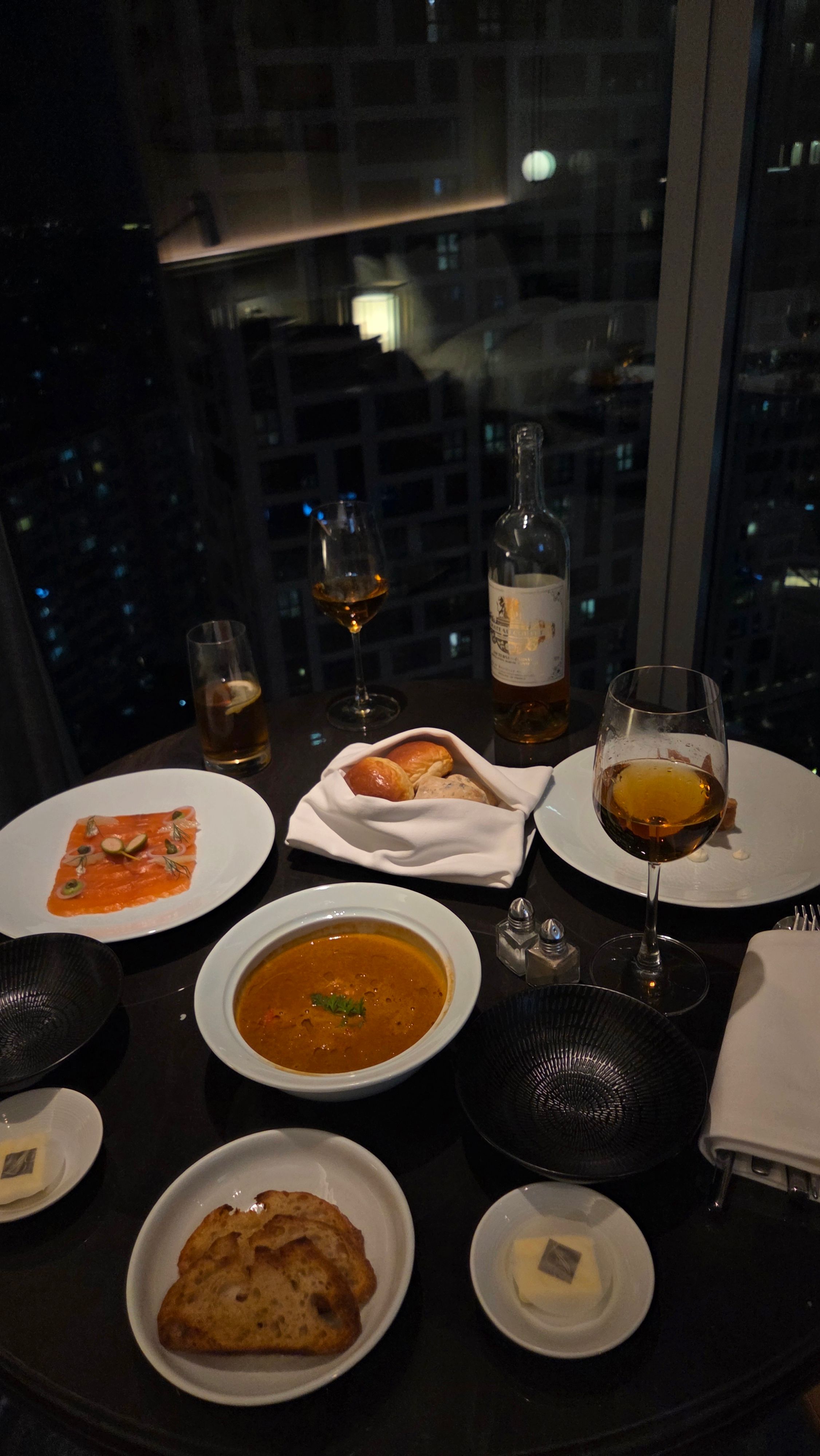 A Seoul hotel table beside a night window with wine, bread, and city reflections.