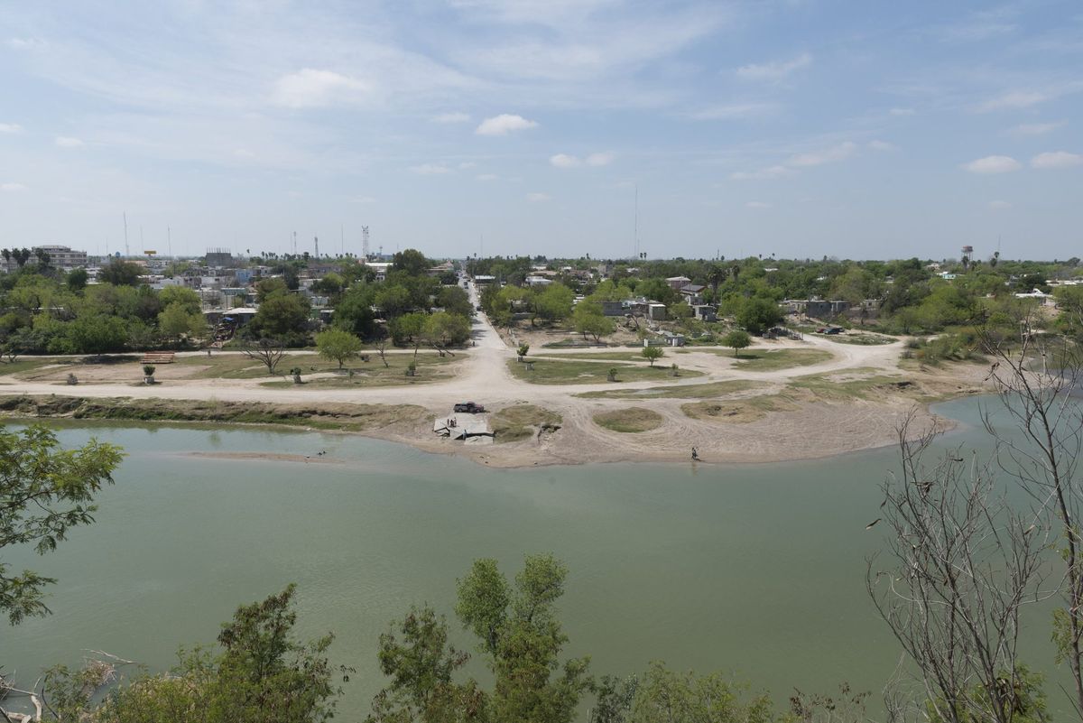 The Rio Grande seen from a bluff, a green river bending below a sandy shore with houses, roads and trees on the Mexican bank beyond.
