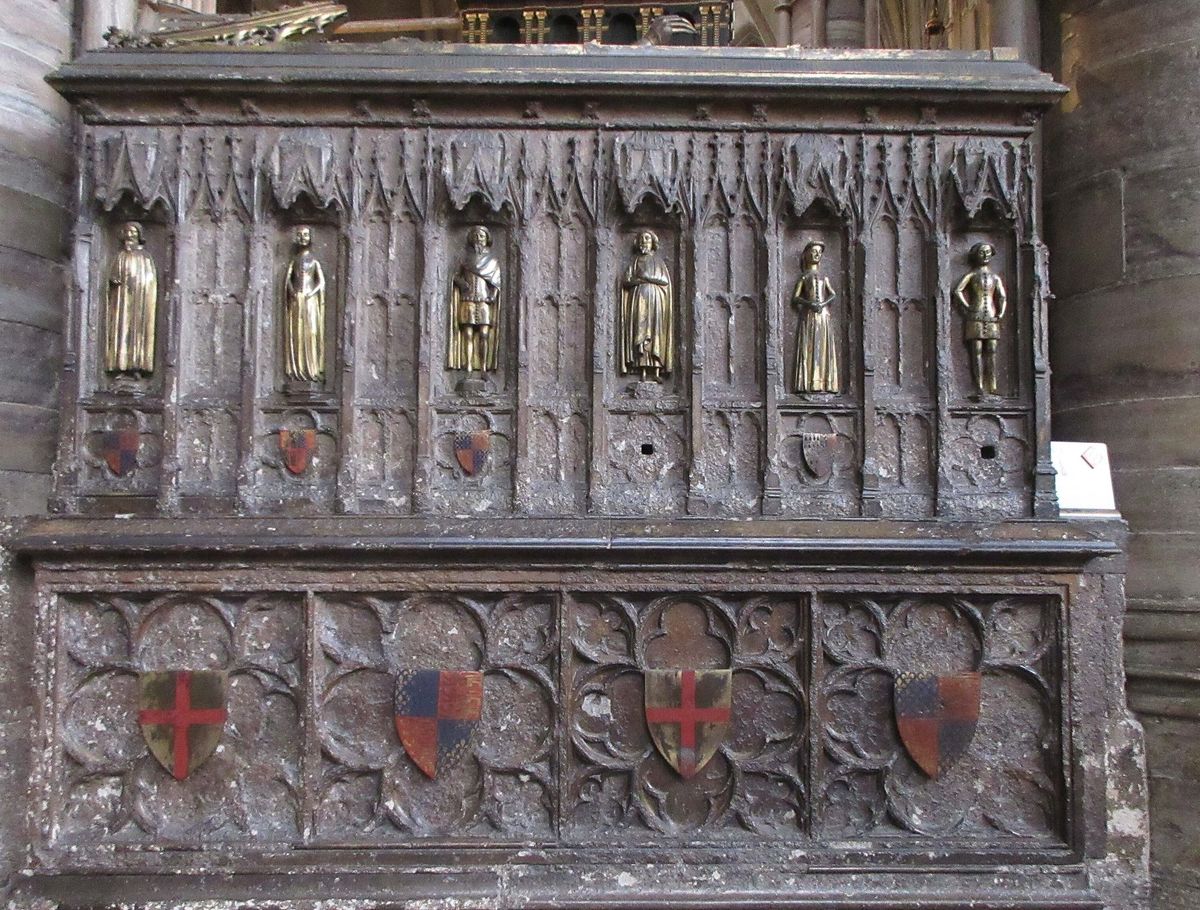 Carved stone side of a medieval tomb with six small gold figures in arched niches above painted shields.