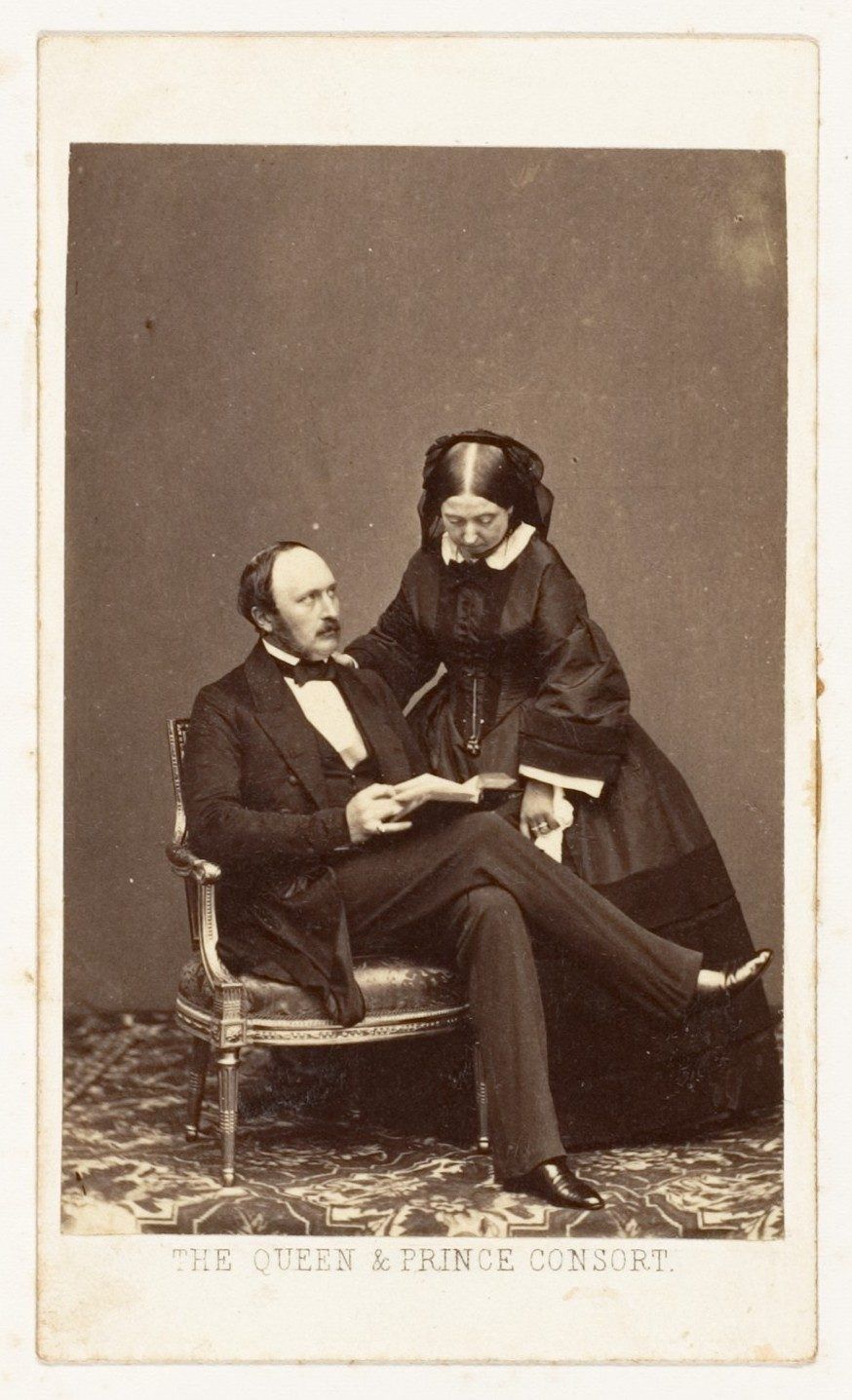 Sepia card photograph: Prince Albert sits holding an open book while Queen Victoria, in a dark dress and white collar, leans over his shoulder.