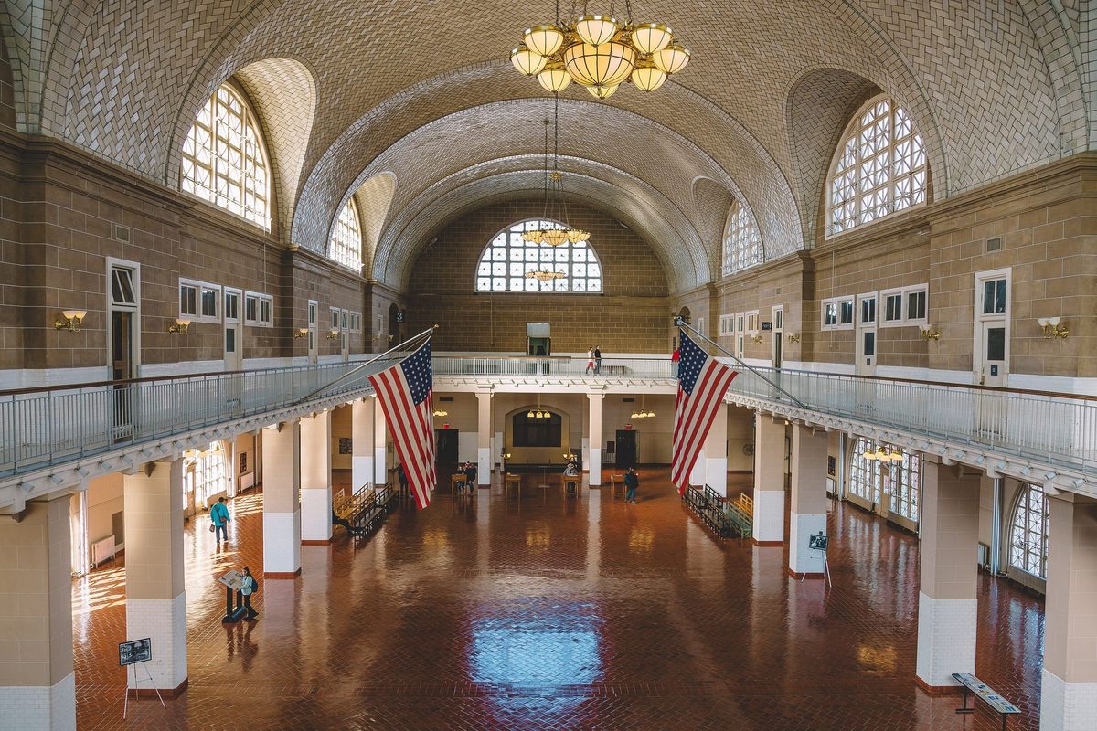 A large vaulted hall with a tiled ceiling, tall arched windows, two hanging American flags and a red tile floor, with a few small visitors in the distance.