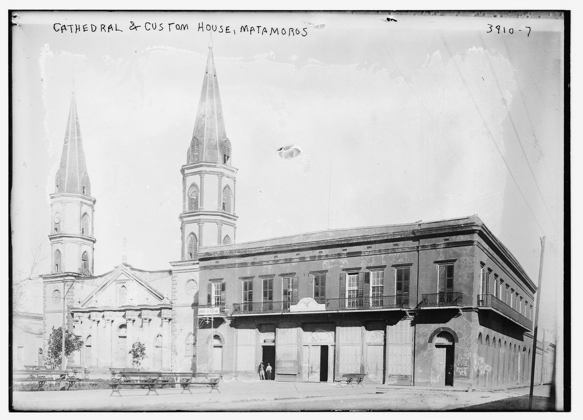 A black-and-white photograph of a church with two tall spires on a plaza beside a long two-story building with balconies, with park benches in the foreground.
