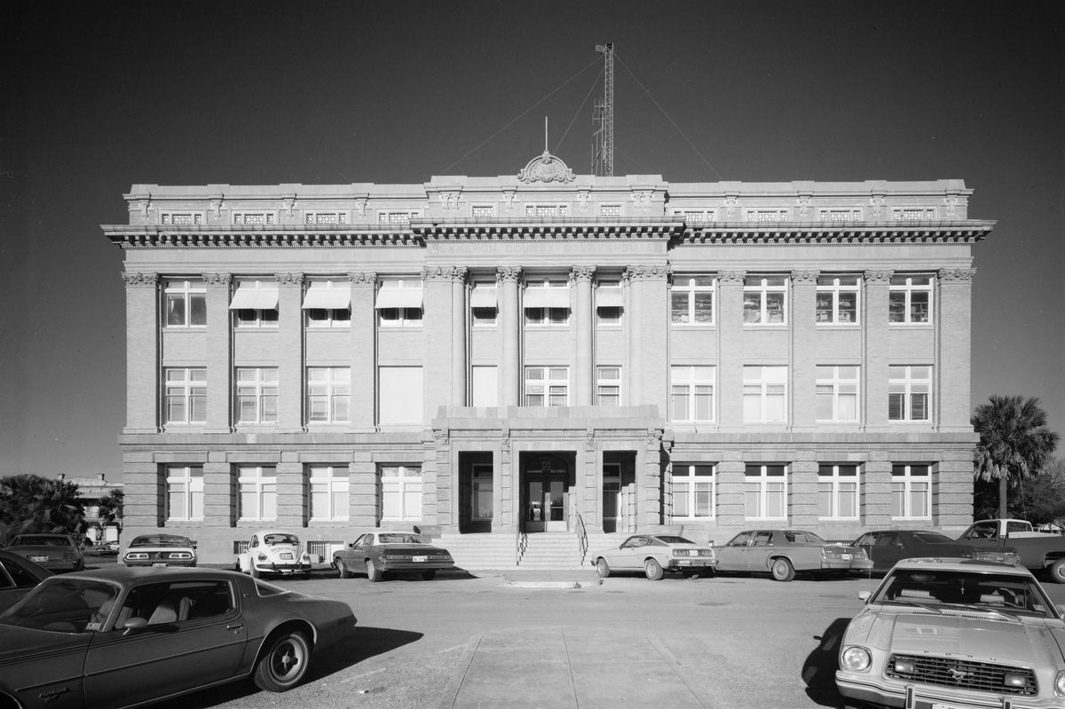 A black-and-white photograph of a three-story brick and stone county courthouse with a columned center entrance and cars parked in front.