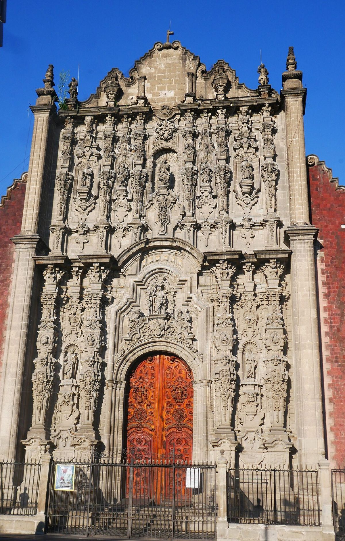 The carved stone facade of the Sagrario Metropolitano in Mexico City, with an ornate orange wooden door below layers of statues and columns.