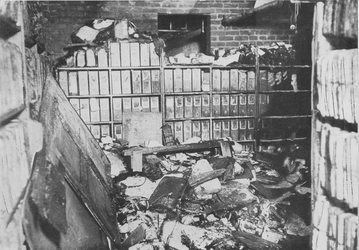 A photograph of charred filing cases, boards and loose papers on shelves and heaped on the floor in a brick-walled room.