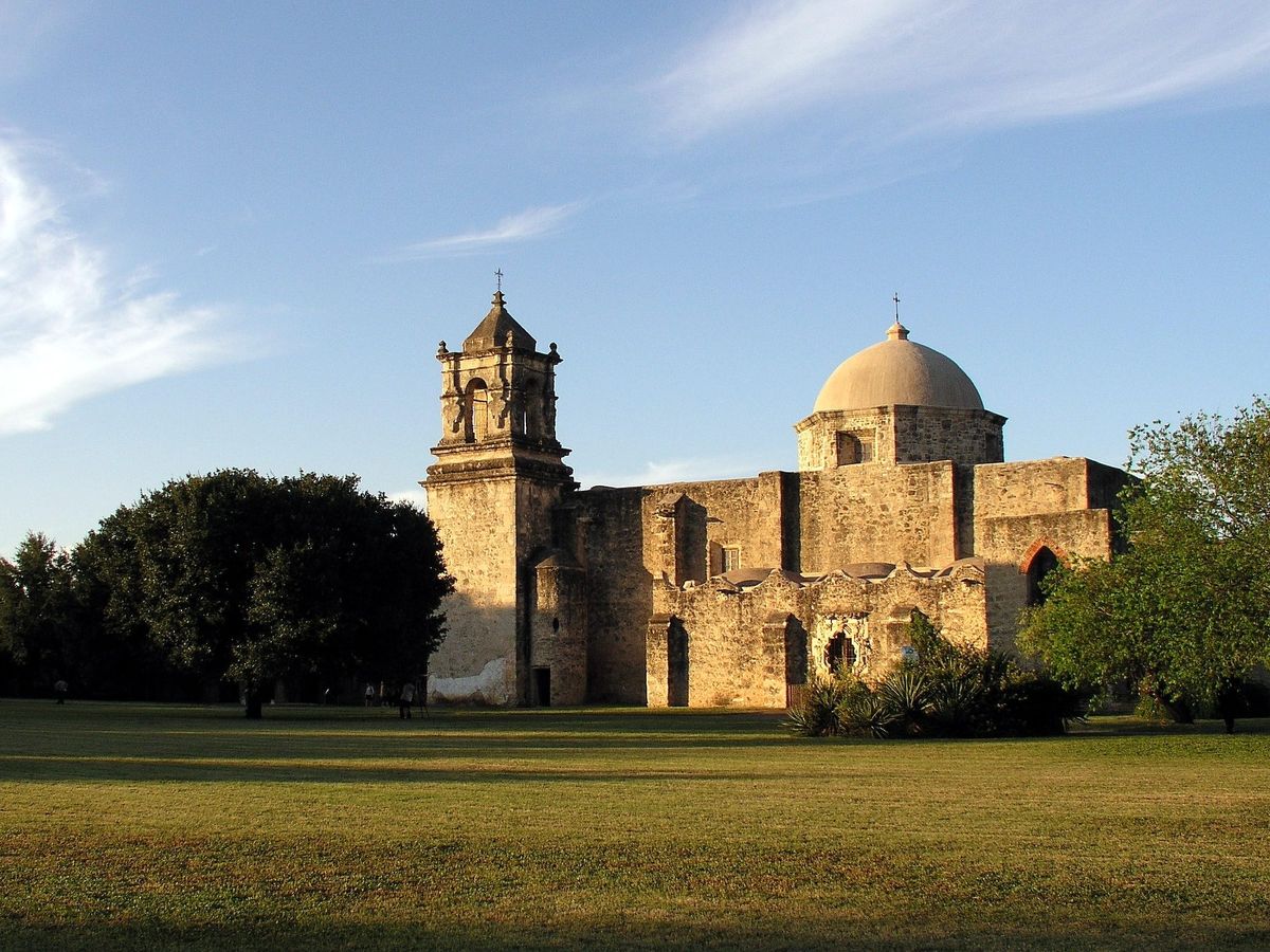 The limestone church of Mission San José in San Antonio, with a bell tower at left and a pale dome at right, seen across a lawn in late sun.