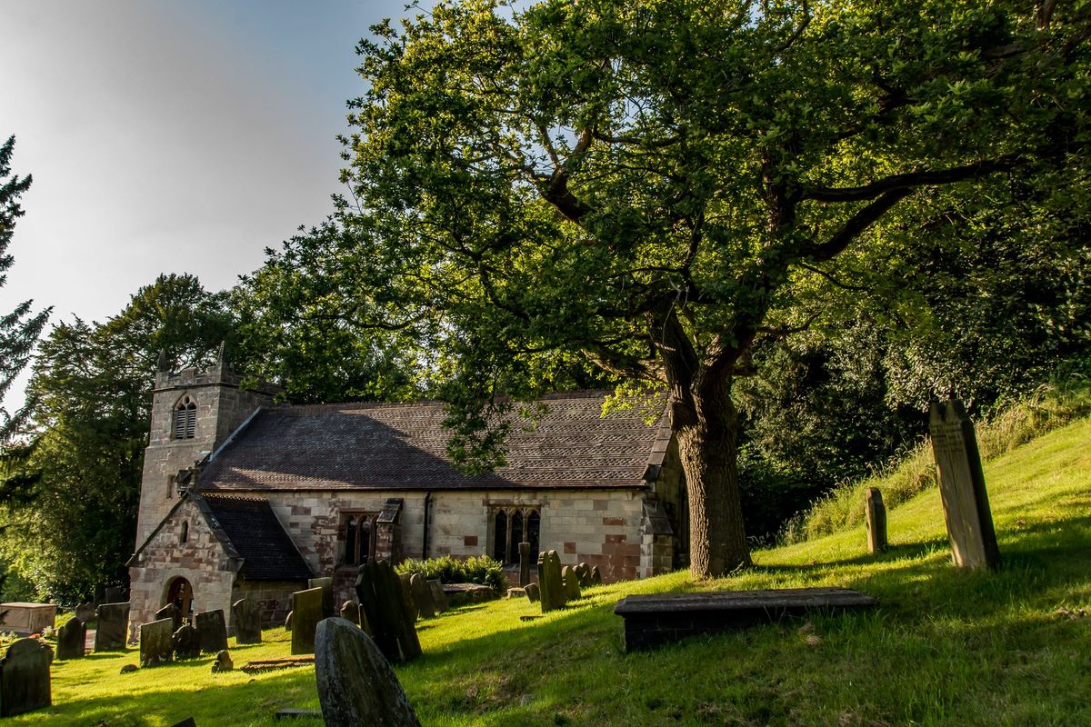 Small stone church with a square tower among trees, old gravestones in the foreground.