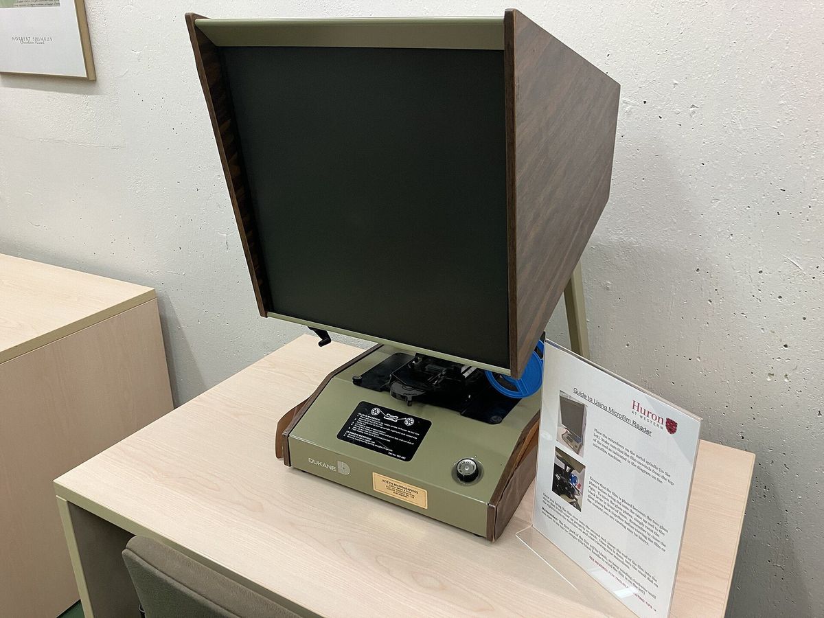 A microfilm reader with a large angled screen and a blue film reel on a pale library desk.