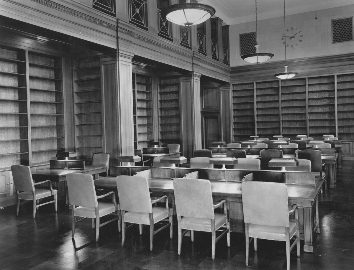 A wood-paneled reading room with long tables, desk lamps, leather armchairs and tall empty bookshelves.
