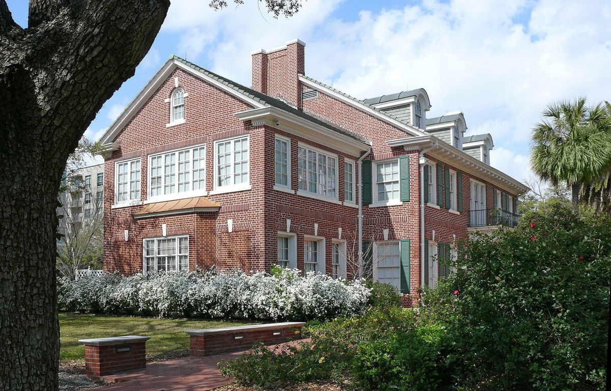 A two-story red brick house with white trim, green shutters and a large live oak.