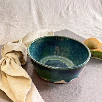Handgjord keramikskål i mörkt jadegrön glasyr. Skålen står på ett grått underlag med en linneservett och kiwi i mjukt naturligt ljus.
