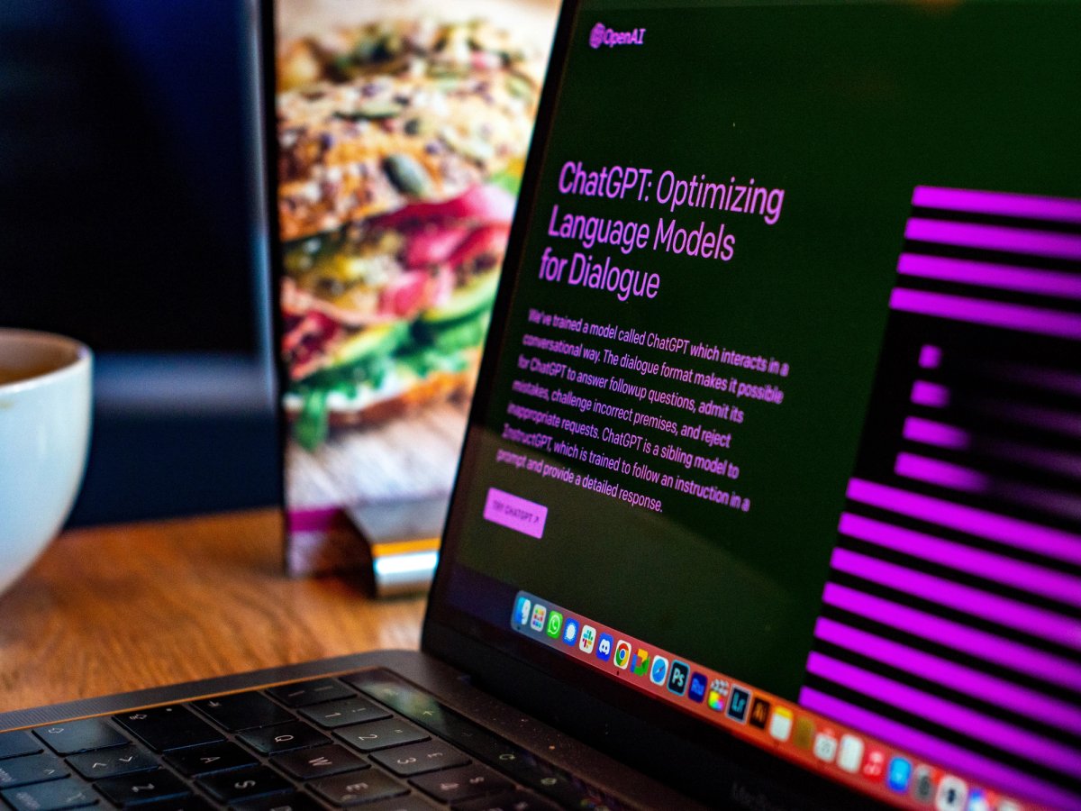 A laptop screen displays the ChatGPT interface with the title “ChatGPT: Optimizing Language Models for Dialogue” in pink text, while a sandwich menu and a coffee cup are visible in the background on a wooden table.