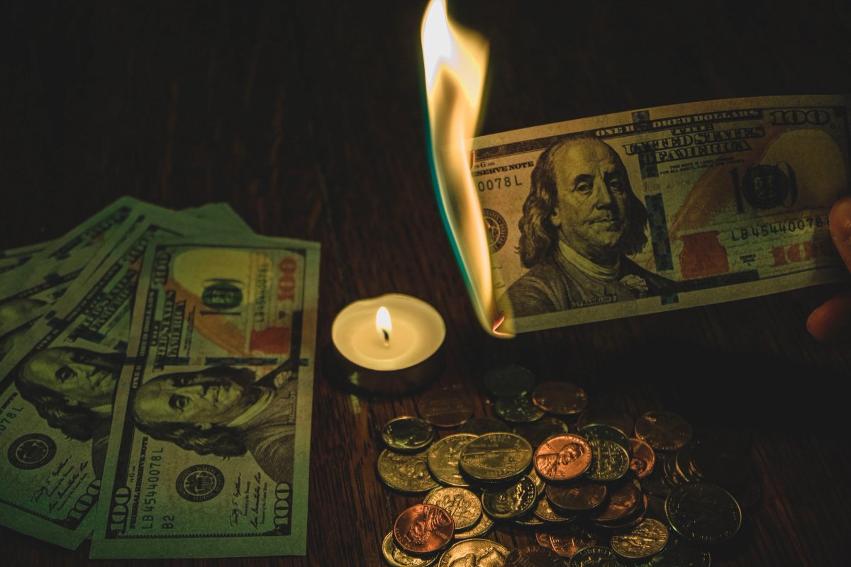 A person burns a $100 US dollar bill next to a small candle, with other hundred-dollar bills and scattered coins on a dark wooden surface, symbolizing wasteful spending or financial loss.