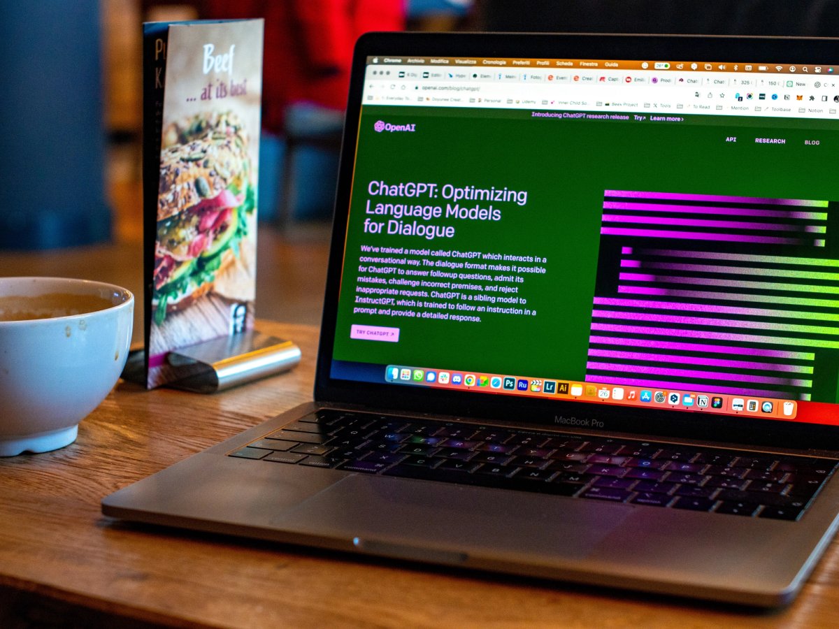 Open laptop on a wooden café table displaying the ChatGPT webpage titled "ChatGPT: Optimizing Language Models for Dialogue," next to a coffee cup and a sandwich menu.