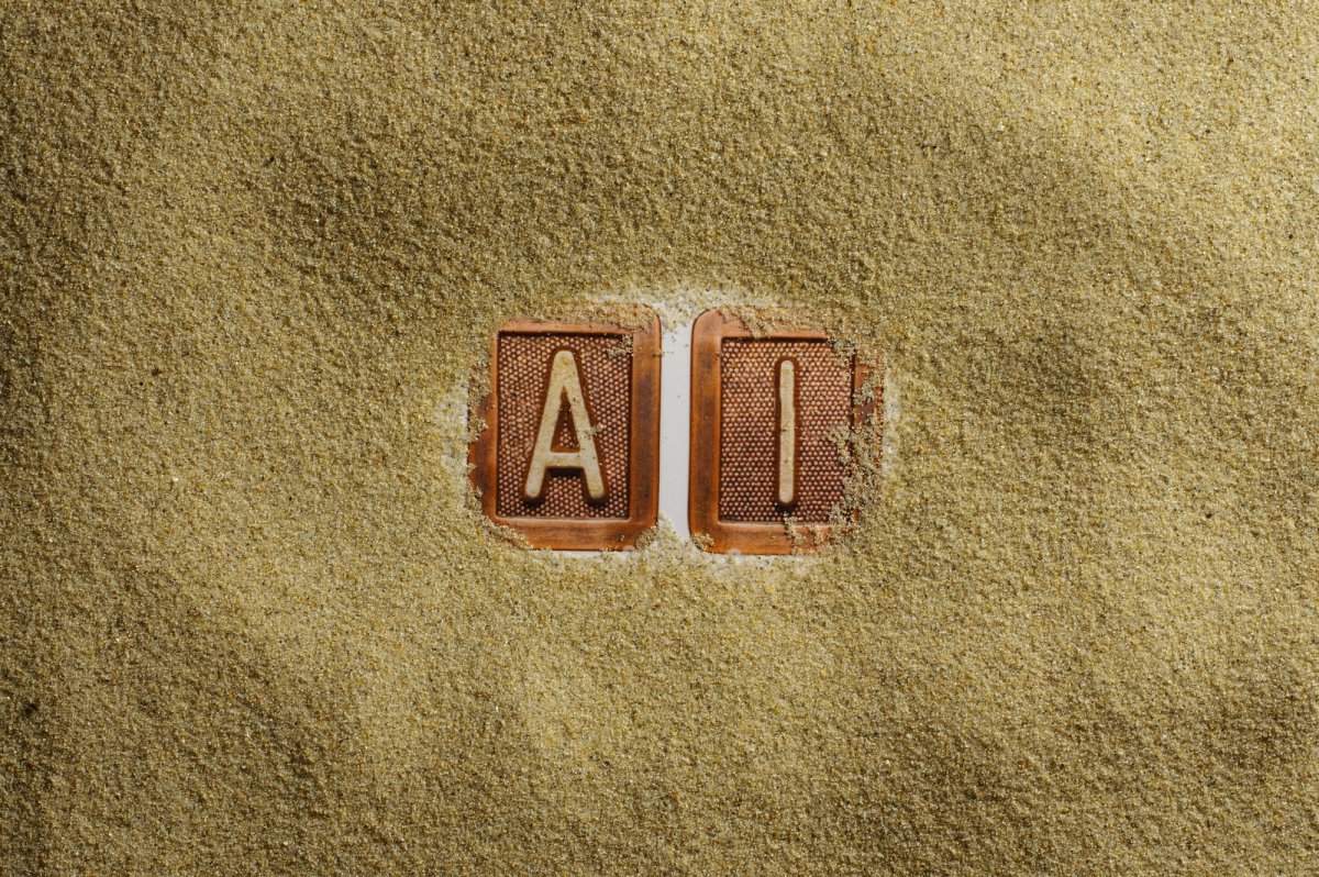 Close-up image of the letters "A" and "I" on brown keyboard-style tiles partially buried in golden sand, symbolizing artificial intelligence emerging or hidden in plain sight.