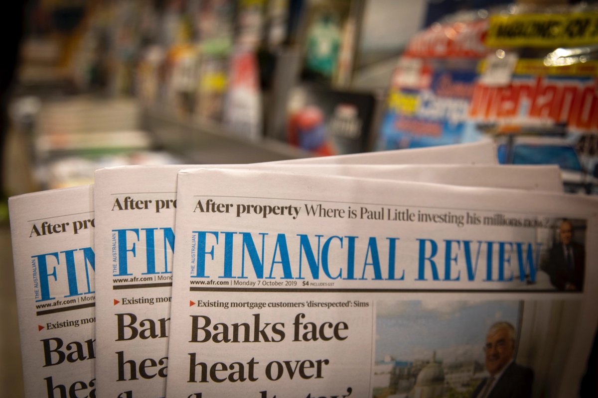 Stack of Australian Financial Review newspapers displayed on a newsstand. The headline reads "Banks face heat over..." with smaller subheadlines above, and various blurred magazines are visible in the background.