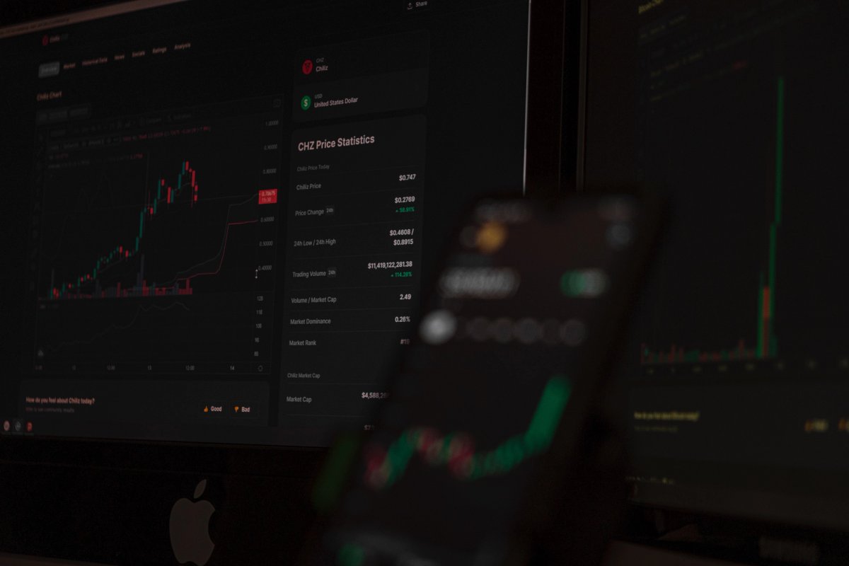 Cryptocurrency trading dashboard displaying price statistics and candlestick chart for Chiliz (CHZ), with a blurred mobile screen in the foreground showing a similar upward trend graph.