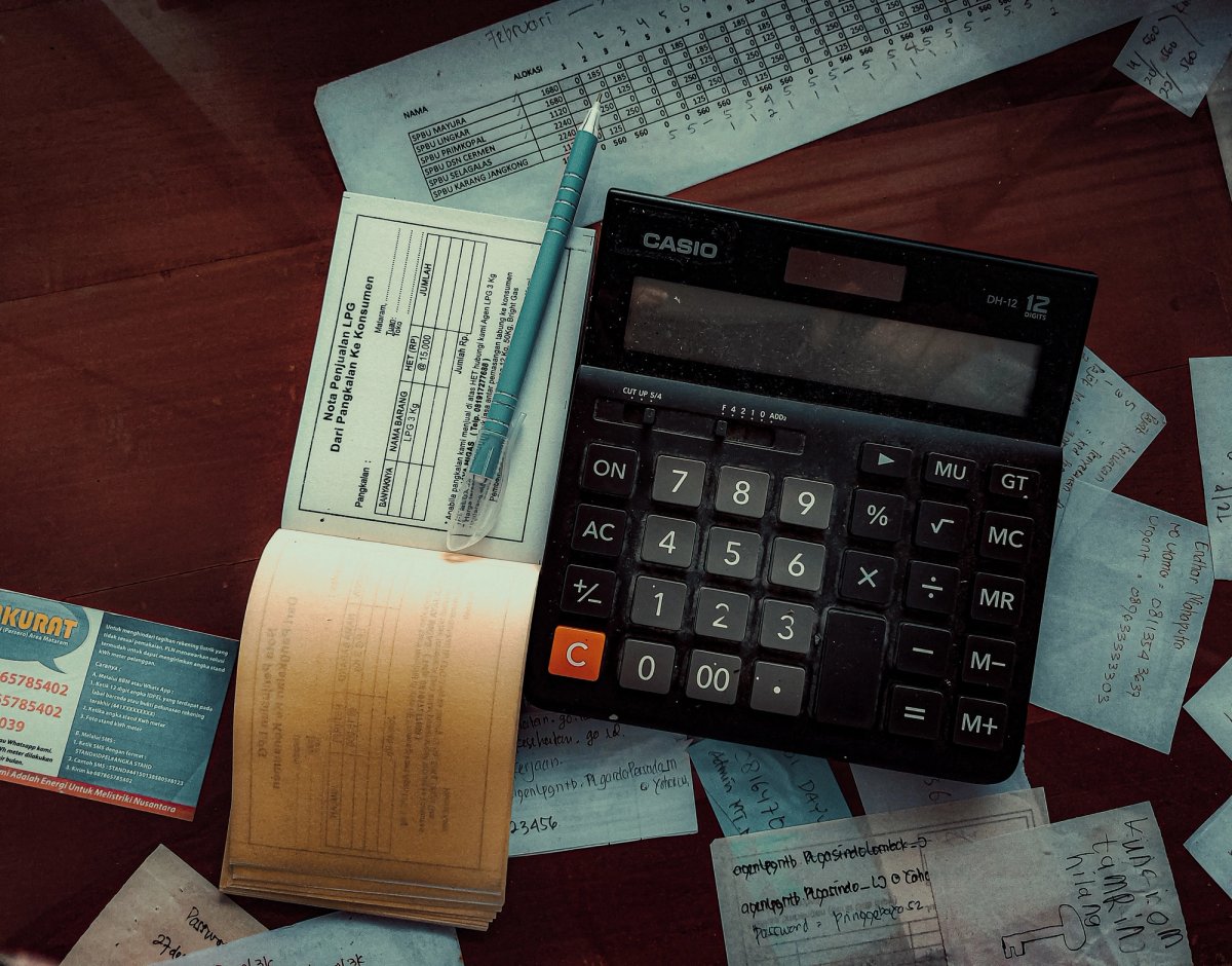 A Casio calculator, pen, and LPG sales receipt book are placed on a desk surrounded by handwritten notes and business cards, suggesting manual financial record-keeping.