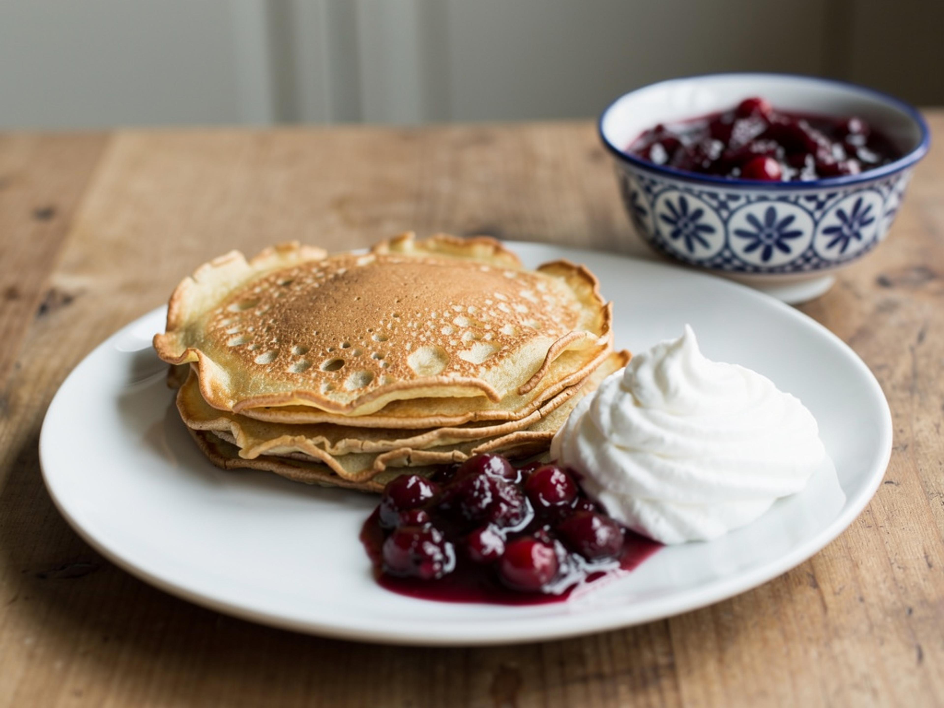 Stack of small thin Swedish pancakes on a white plate with lingonberry preserves and whipped cream