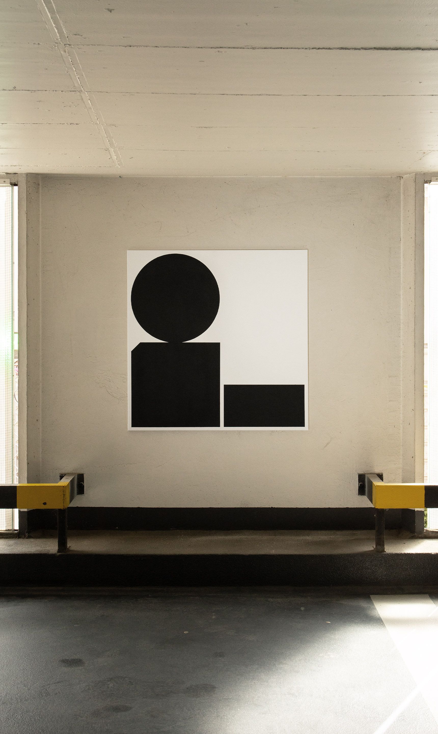 Black and white abstract acrylic painting on canvas with geometric shapes — two rectangles and a balancing circle — displayed in a parking garage.