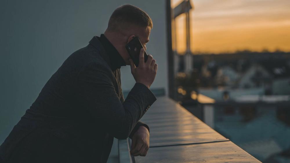 A man on a phone call, leaning on a rooftop railing at sunset.