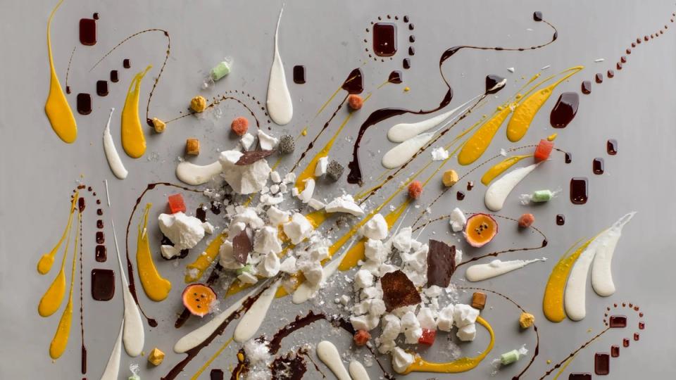 A dessert painted across a grey table: streaks of mango and chocolate, broken meringue and passion fruit.