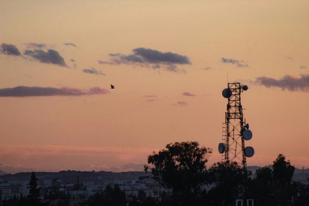 A telecommunications tower hung with satellite dishes, silhouetted against a pink and orange sunset over a city.