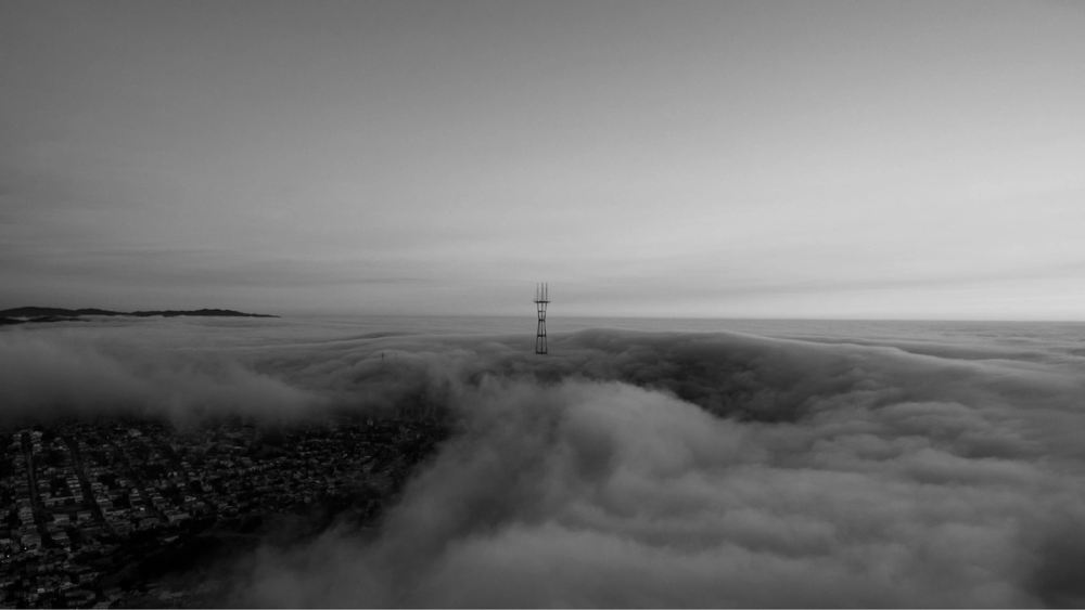 A radio tower rising above a thick bank of fog rolling over a city, in black and white.