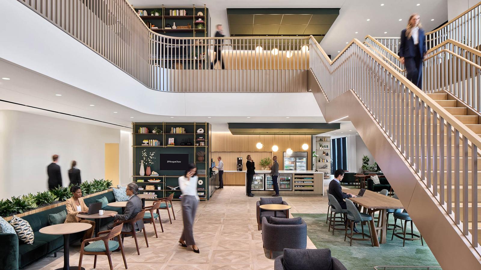 A double-height lounge with a curved balcony and bookshelves, people working at tables below.