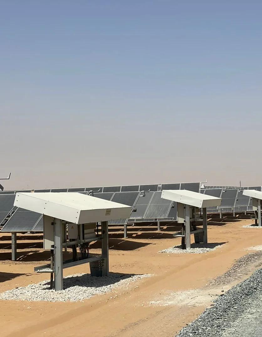 Electrical inverter enclosures with protective sunshades in the foreground of a desert solar farm with panel arrays extending to the horizon