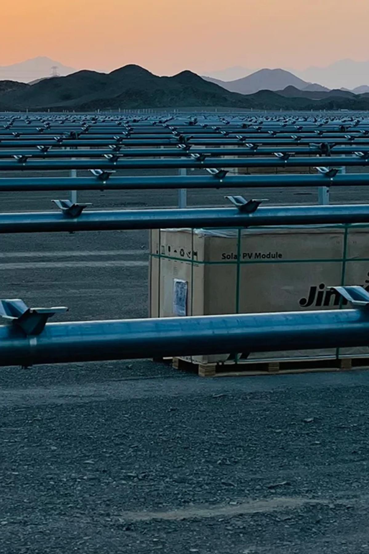 Solar PV modules on pallets at a construction site with mounting structures and mountains at dusk