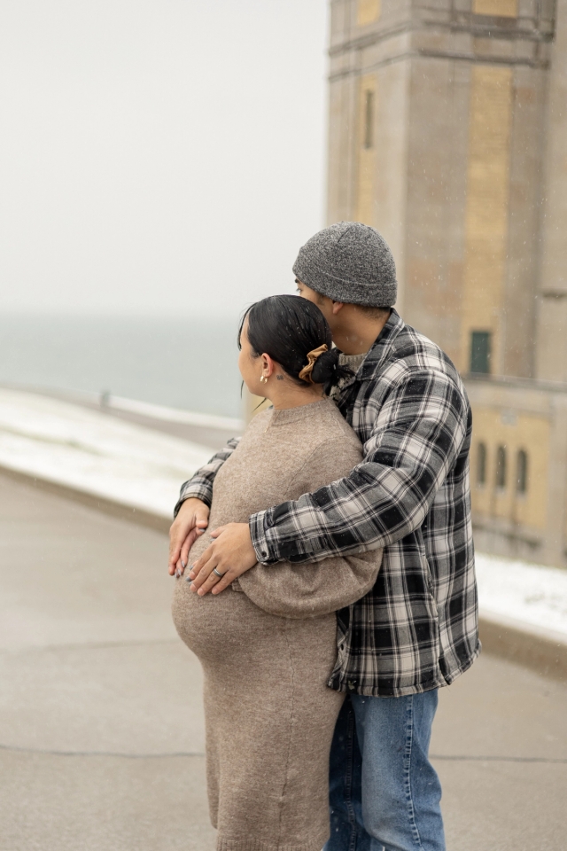 Scarborough maternity photography featuring a glowing mother-to-be and her husband embracing her in a hug from behind under soft, natural winter light at the R.C. Harris Water Treatment Plant