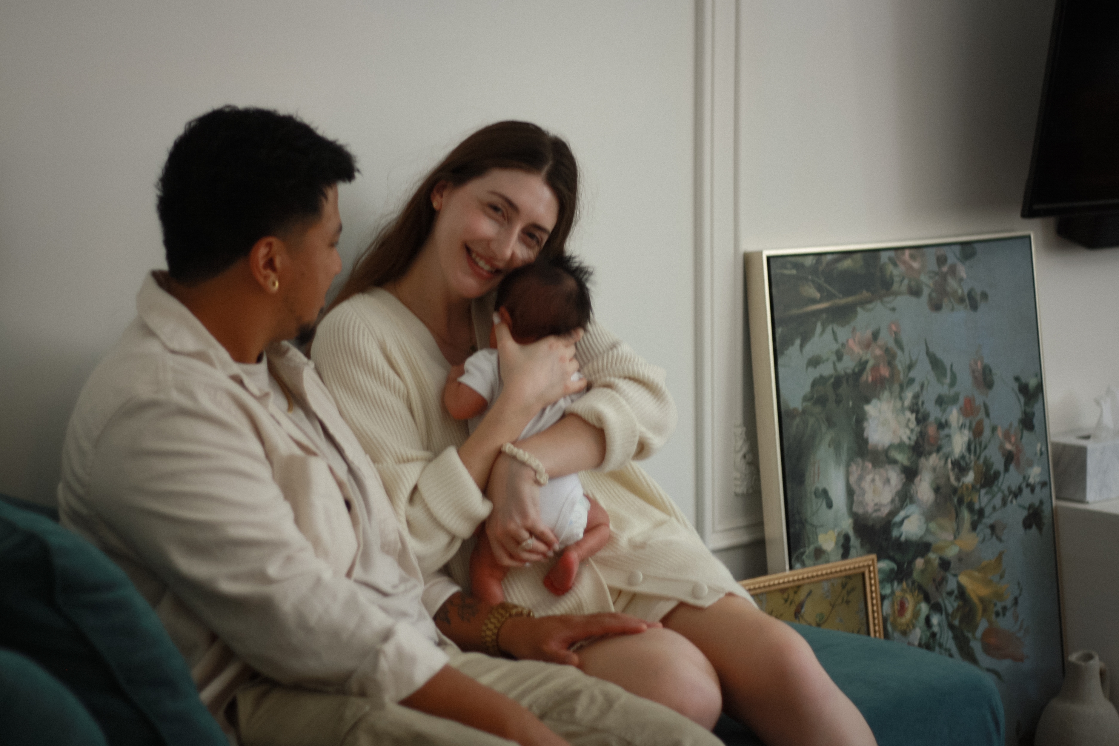 Couple sitting on couch, dad looking at mom while she smiles at camera hugging baby close to her chest, picture frame with a floral paining in background - Newborn Photography