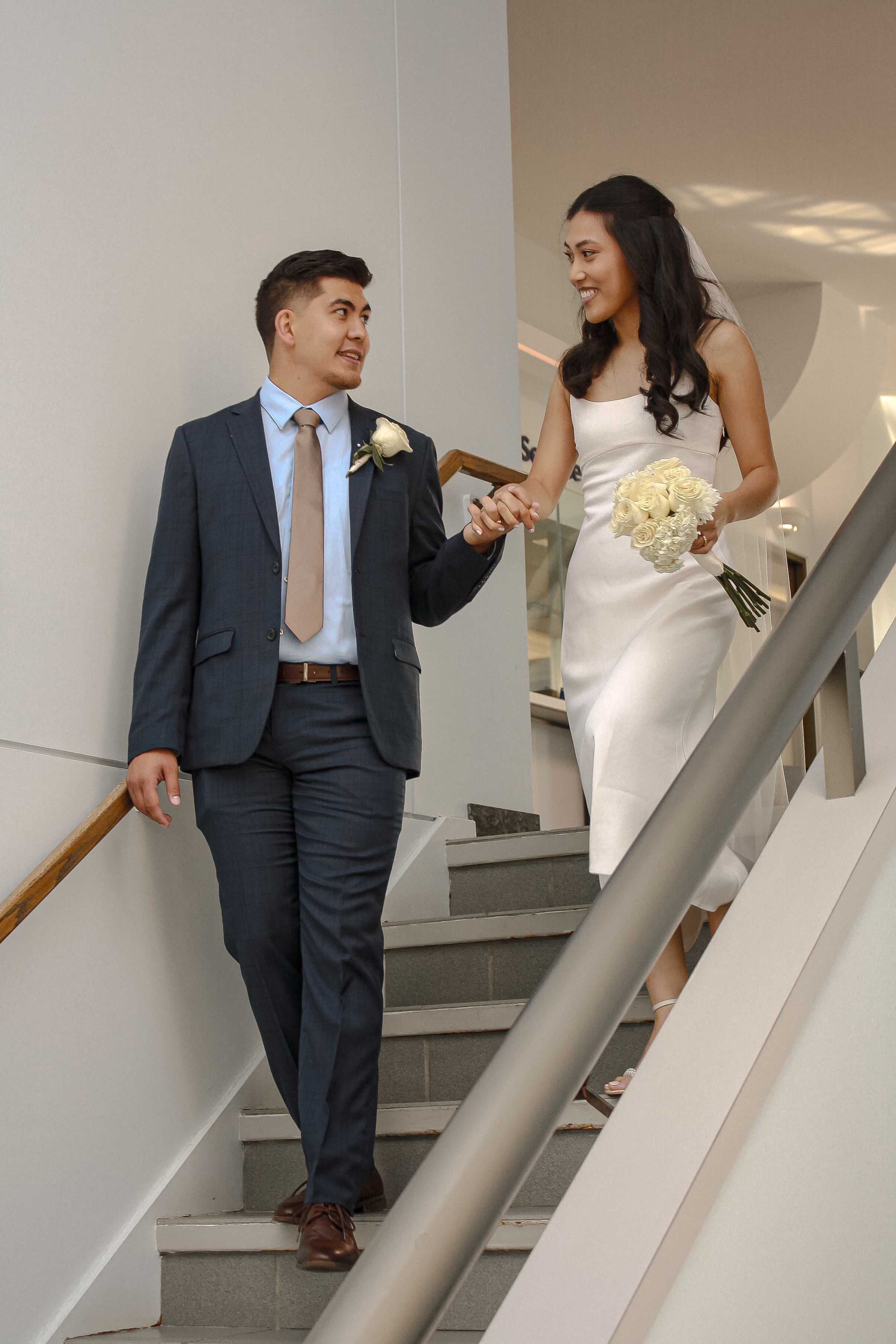 Bride and Groom walking down a set of stairs holding hands and smiling at each other, in a courthouse, bride is holding a bouquet of white roses - Wedding Photography