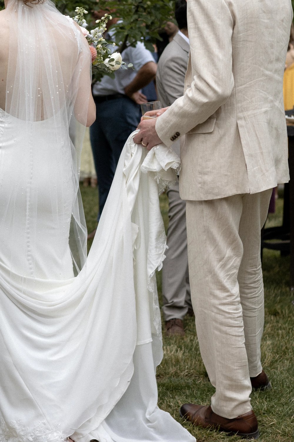 groom holding bride's wedding dress train while interacting with guests at outdoor summer wedding