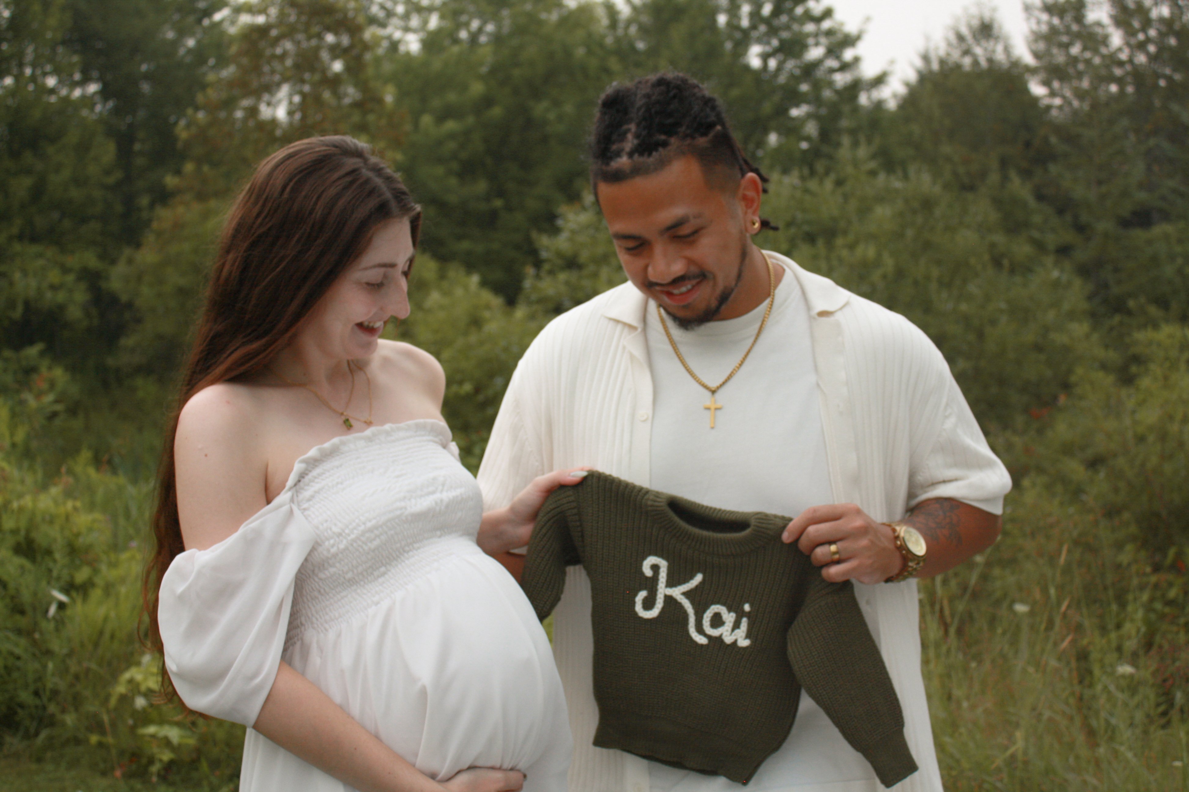 couple dressed in white standind in front of trees holding baby bump and a green knit sweater with the name 'Kai' stitched on it, smiling - Newmarket Maternity Photography