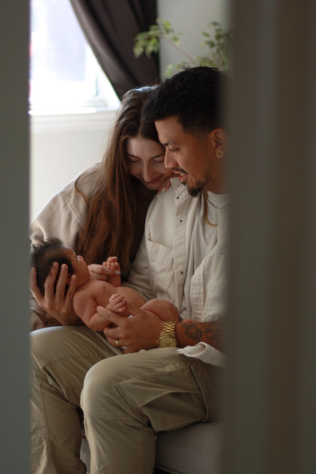 parents sitting on bench at end of bed looking lovingly at baby while dad cradles his head - newborn photography