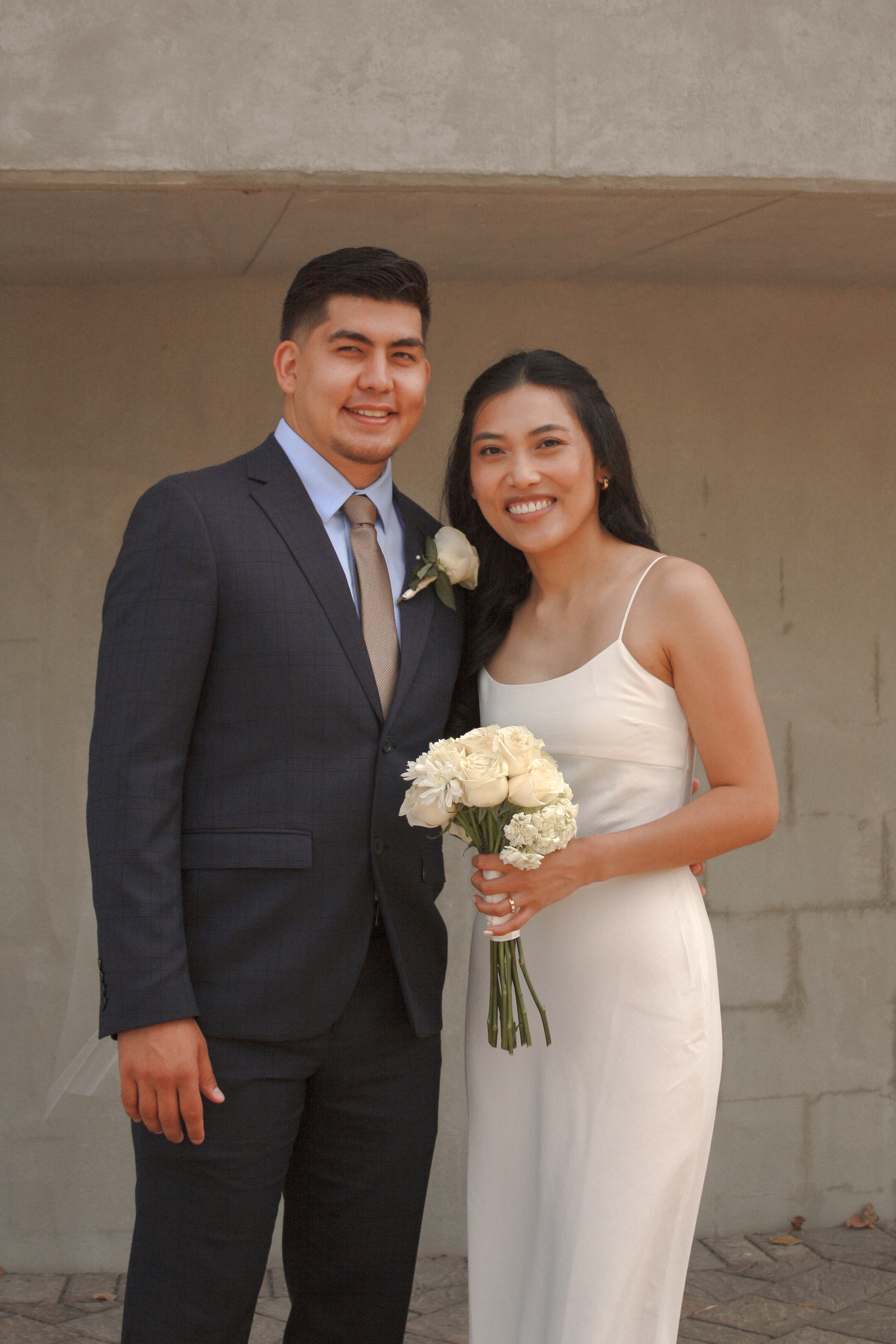 bride and groom standing beside each other smiling at camera while bride holds a bouquet of white roses with long stems - Wedding Photography