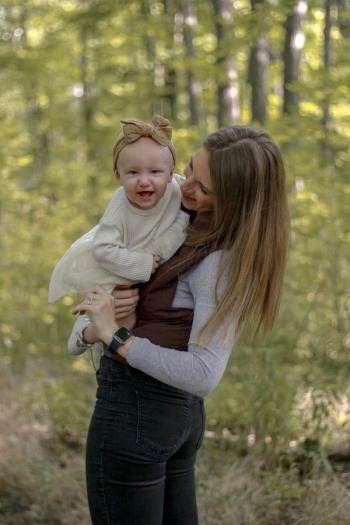 mom standing sideways holding baby girl in white dress and a bow on her head, in the forest - Family Photography