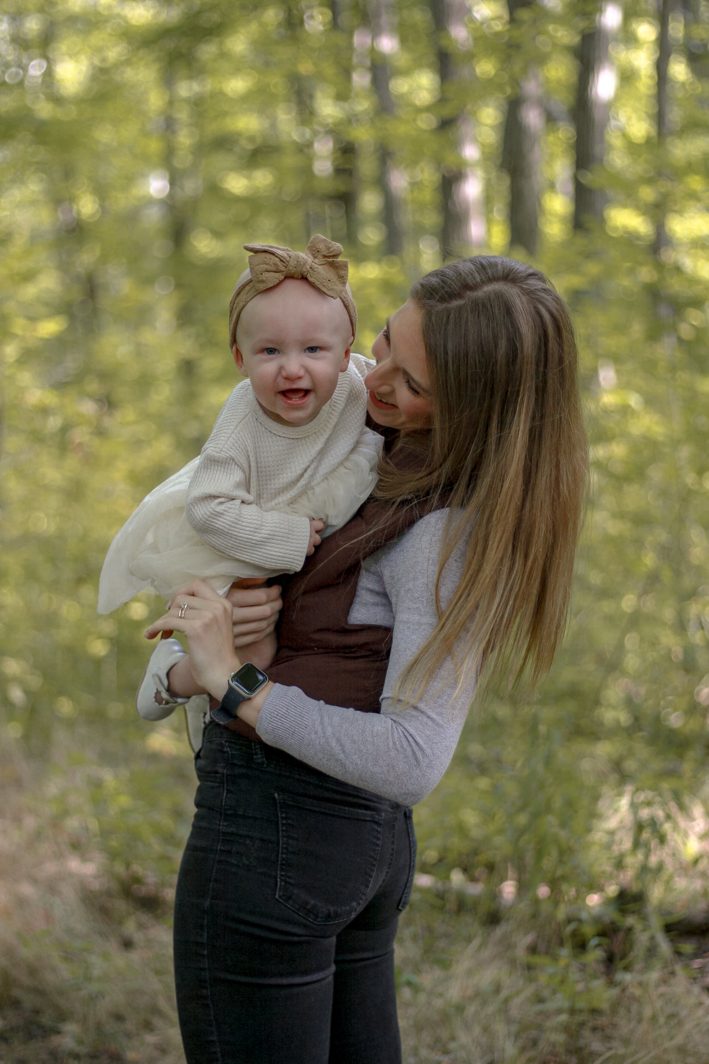 mom standing sideways holding baby girl in white dress and a bow on her head, in the forest - Newmarket Family Photography