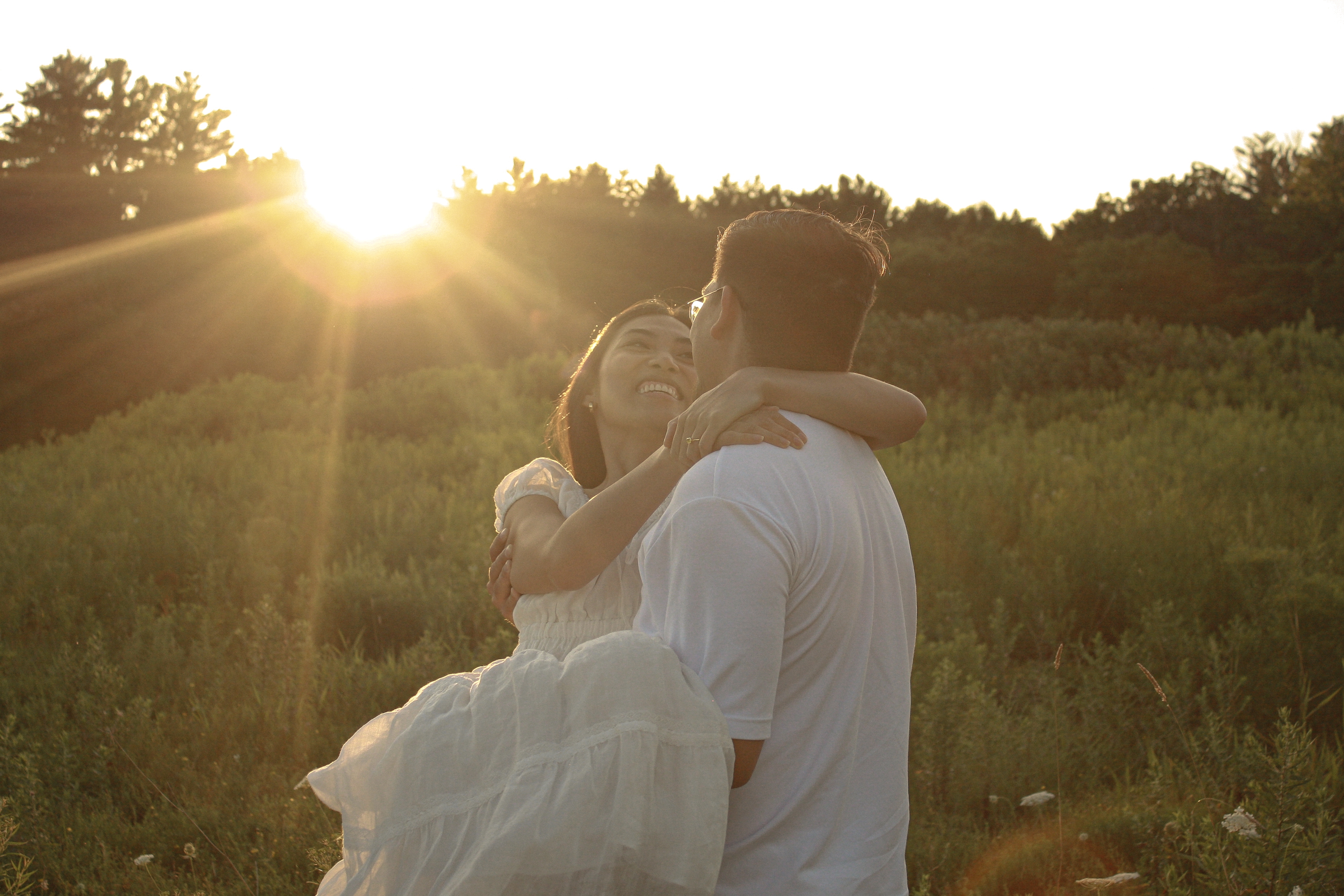 Couple in a grass field, sun is setting and glaring through the trees, man in white shirt is holding his fiancée with his back to camera, she is smiling at him - Engagement Photography