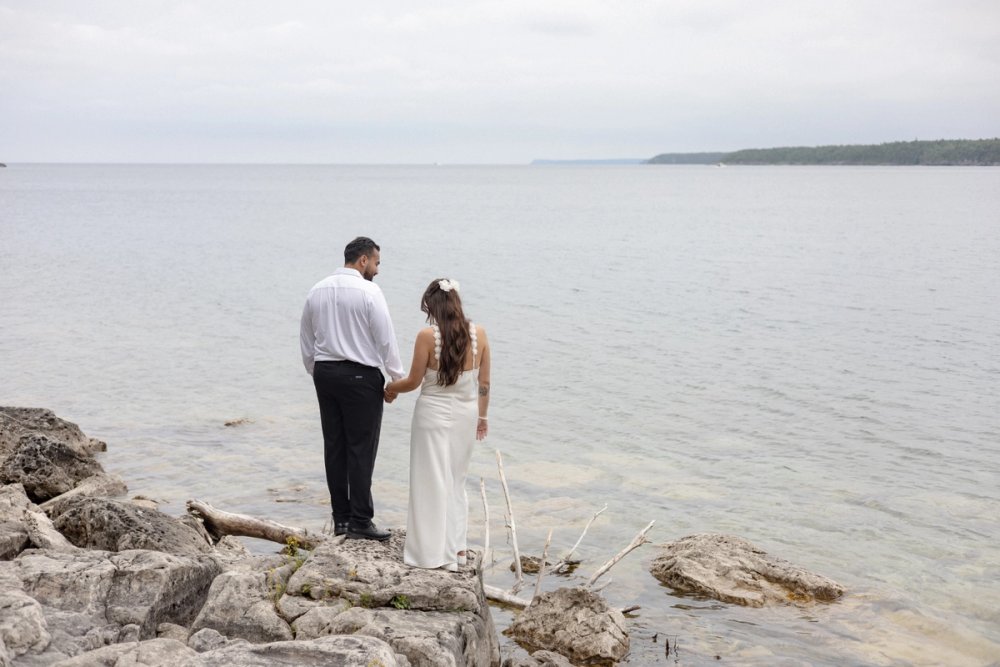 Bride and groom standing by rocks along a shoreline - Tobermory Elopement Photography