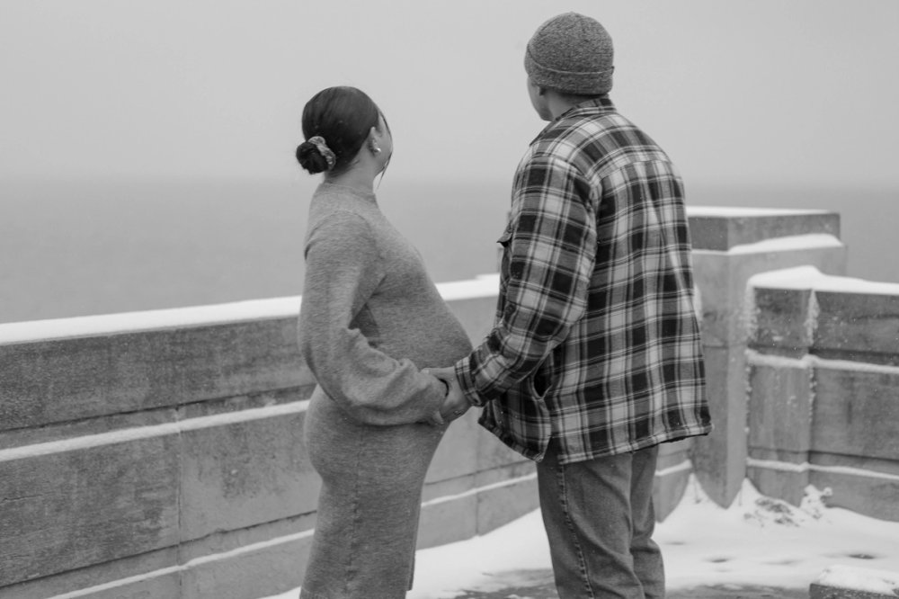 Expecting first-time parents holding hands during a winter maternity photoshoot in Scarborough, Ontario while looking out at lake. - Newmarket Maternity photography