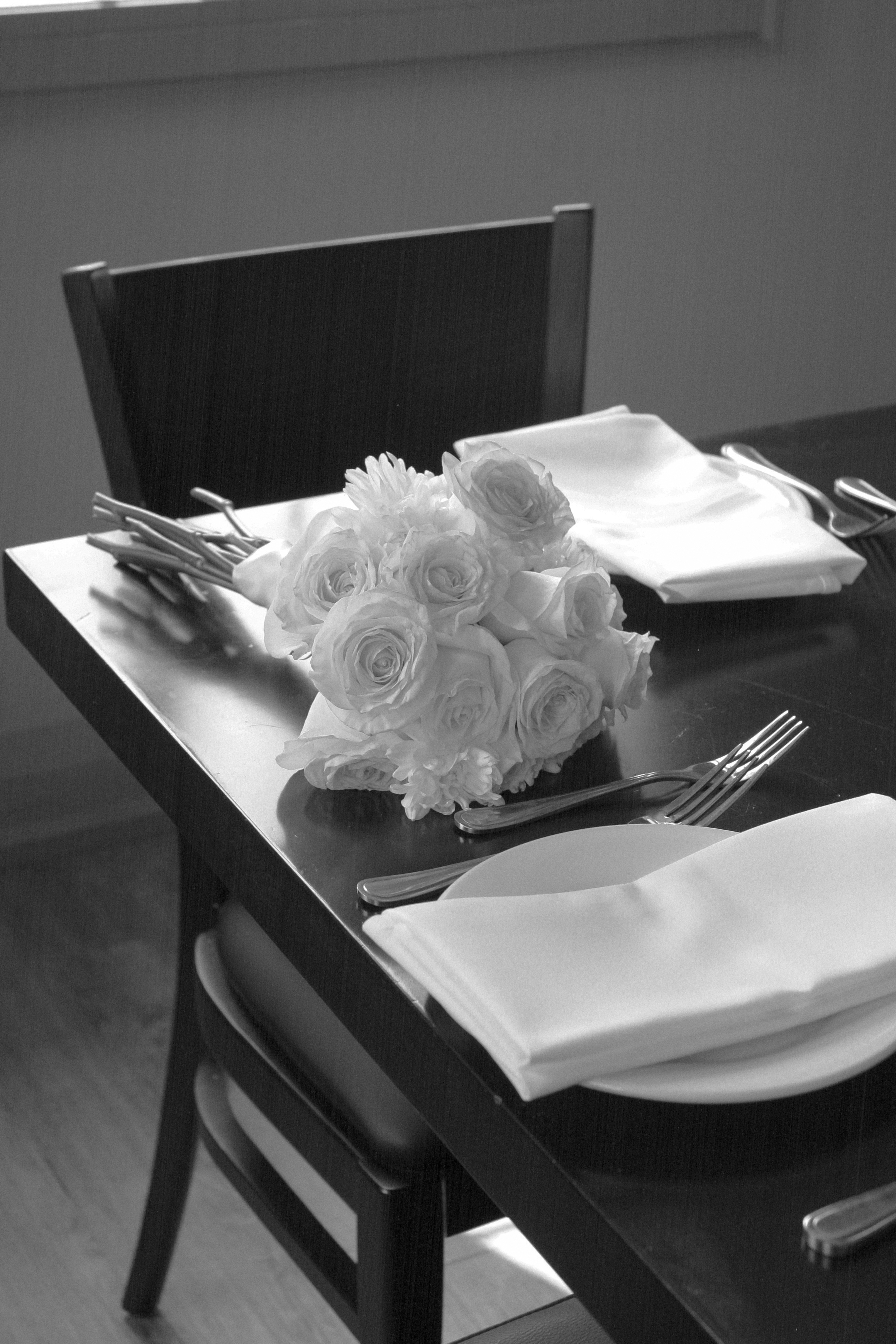 black and white photo of bouquet of white roses on table corner with white plates and napkins set up and chair behind table and flowers - Wedding Photography