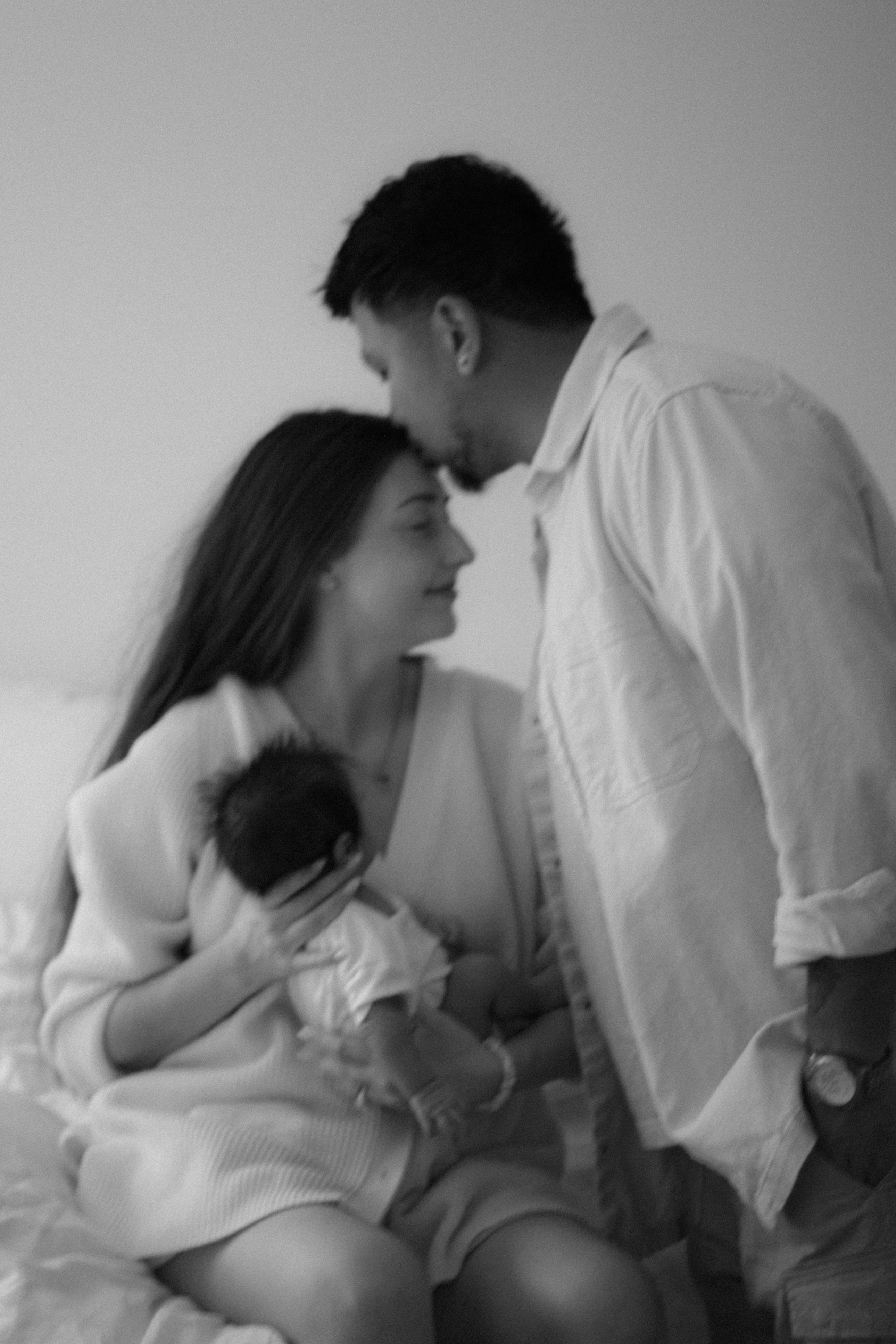 black and white photo of mom sitting on bed holding newborn baby in arms while dad stands next to bed kissing moms forehead - Newmarket Newborn Photography