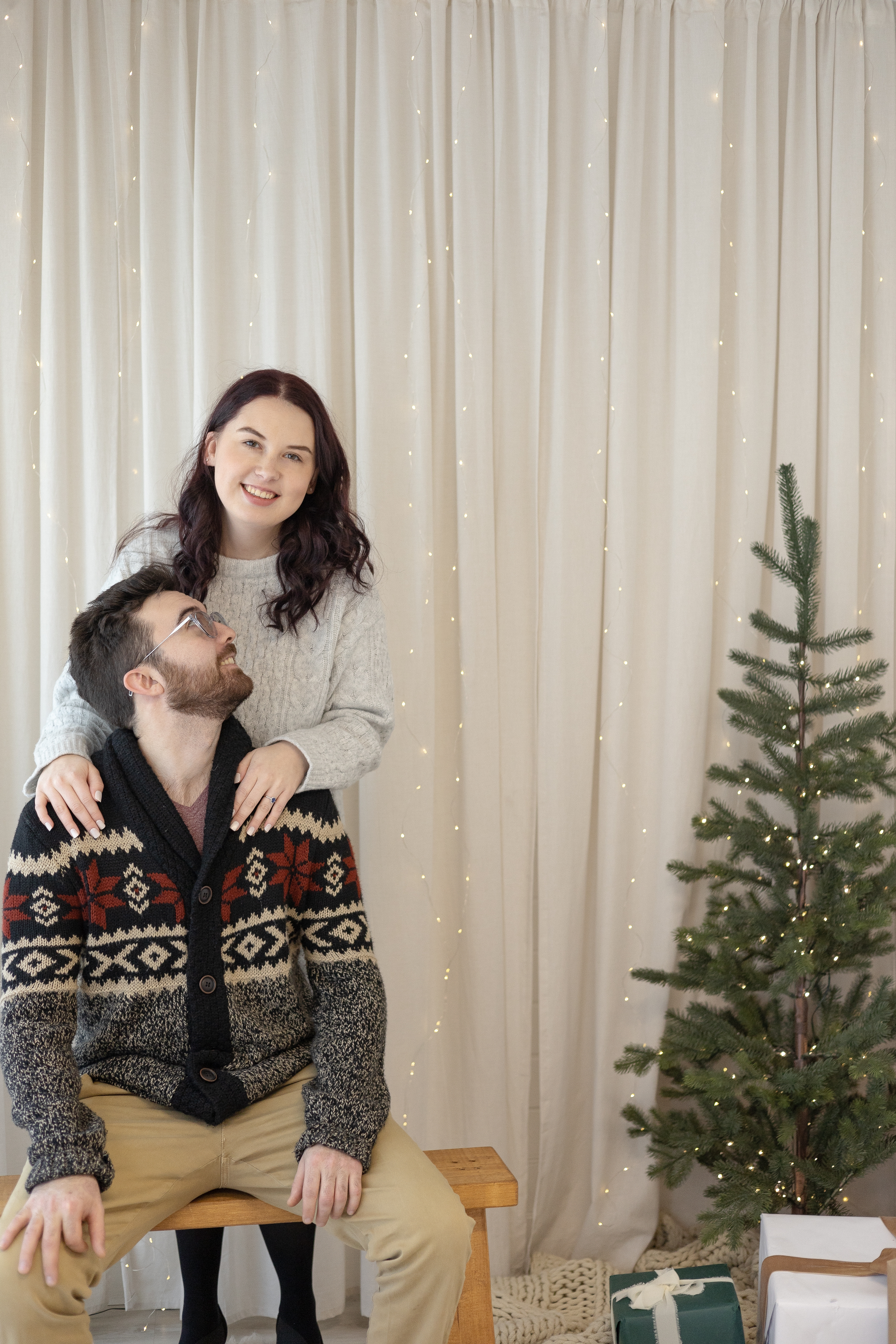 Christmas Studio set up with engaged couple, sitting on a wooden bench with a white backdrop and mini Christmas tree