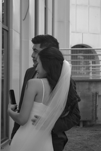 candid photo of bride holding phone taking a photo of a window reflection while groom has his hand on her lower back smiling and both looking left - Wedding Photography