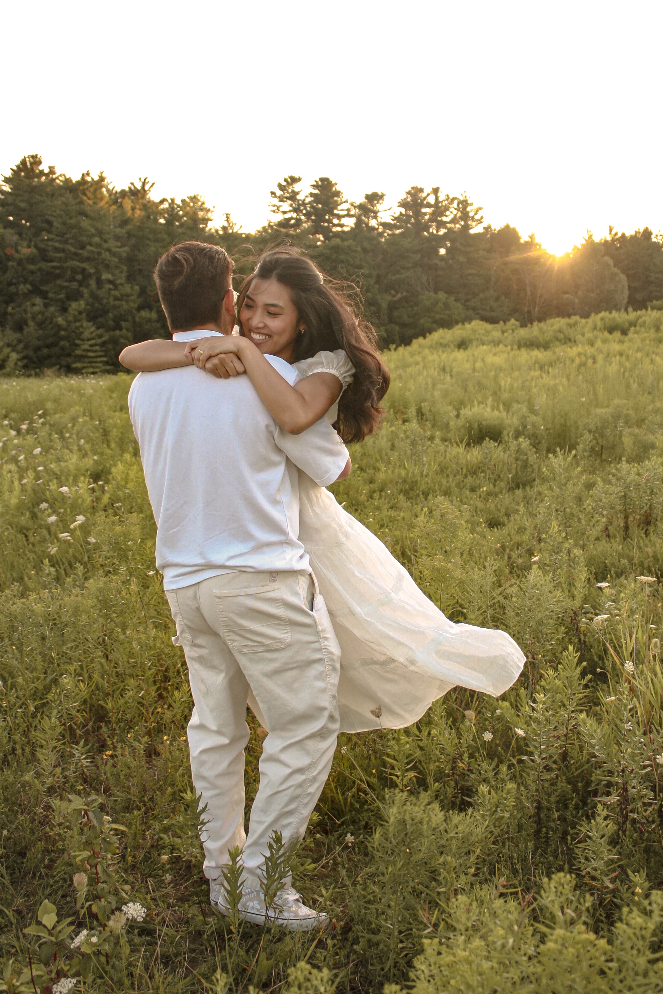 couple in grass field wearing all white outfits at sunset, man is spinning his fiancée and she has her arms around his neck, dress is flowing - Engagement Photography